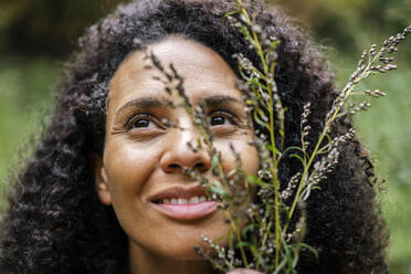 Lächelnde Frau, die eine Pflanze hält, während sie im Wald steht - AKLF00015