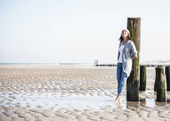 Junge Frau mit geschlossenen Augen lehnt an einem Holzpfosten am Strand - UUF22680