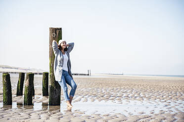 Smiling woman with hands behind head leaning on wood post at beach - UUF22681