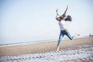 Cheerful young woman with arms raised jumping over wet sand at beach - UUF22682