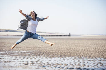 Fröhliche junge Frau, die an einem sonnigen Tag über nassen Sand springt - UUF22685