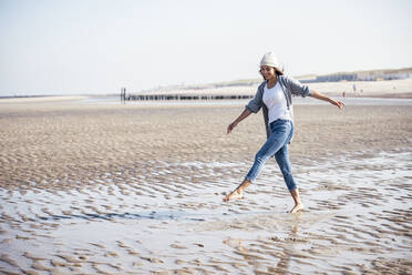 Carefree young woman in knit hat walking on wet sand at beach - UUF22686