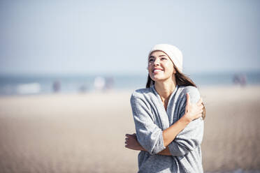 Lächelnde Frau, die sich selbst umarmt, während sie am Strand an einem sonnigen Tag steht - UUF22689