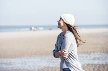 Happy young woman in knit hat hugging self while standing at beach - UUF22690