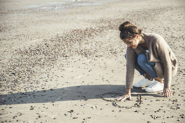 Young woman keeping hand between heart drawn on sand at beach - UUF22714