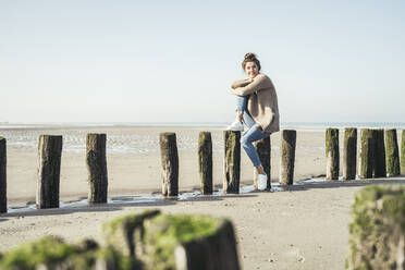 Young woman day dreaming while sitting on wooden post at beach during weekend - UUF22715