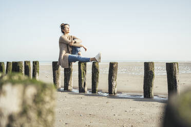 Schöne Frau mit geschlossenen Augen sitzt auf Holzpfosten am Strand während des sonnigen Tages - UUF22716
