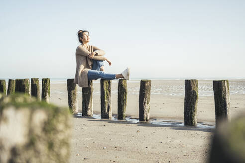 Schöne Frau mit geschlossenen Augen sitzt auf Holzpfosten am Strand während des sonnigen Tages - UUF22716