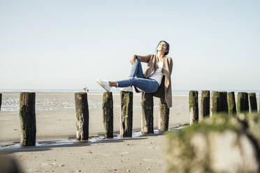 Young woman in cardigan sweater with eyes closed sitting on wood post at beach - UUF22717