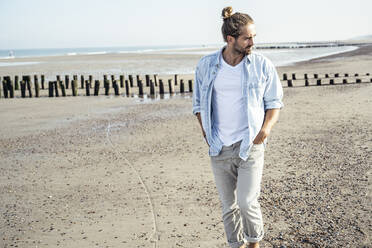 Gut aussehender junger Mann mit Händen in den Taschen, der beim Spazierengehen am Strand wegschaut - UUF22721