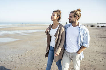 Smiling girlfriend and boyfriend looking away while walking on beach during weekend - UUF22725