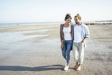 Junges Paar mit Händen in den Taschen bei einem Spaziergang am Strand während eines sonnigen Tages - UUF22726