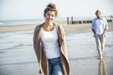 Young couple with hands in pockets walking on beach during sunny day - UUF22729