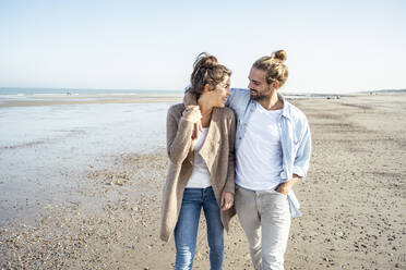 Young smiling couple looking at each other while walking on beach during weekend - UUF22741