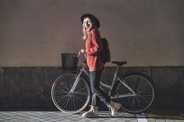 Young woman wearing hat walking with bicycle on footpath - JSMF01894