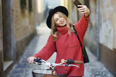 Lächelnde Frau, die ein Selfie mit ihrem Mobiltelefon macht, während sie mit dem Fahrrad auf dem Gehweg steht - JSMF01906