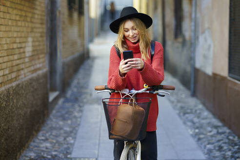 Blonde Frau benutzt ihr Smartphone, während sie mit dem Fahrrad auf dem Gehweg steht - JSMF01907