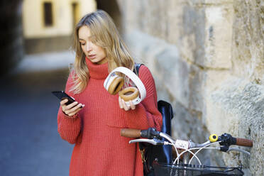 Blonde Frau mit Kopfhörern, die ein Mobiltelefon benutzt, während sie am Fahrrad steht - JSMF01914