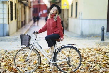 Blond woman wearing hat standing with bicycle on dry leaves over footpath - JSMF01923