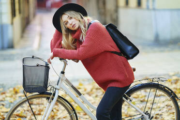 Blonde Frau mit Rucksack lehnt mit dem Fahrrad auf dem Fußweg stehend - JSMF01926