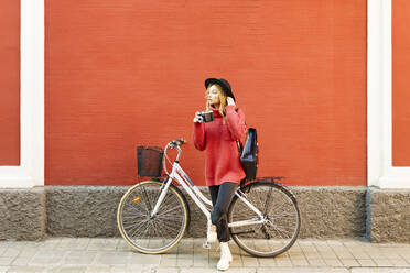 Junge Frau mit Hut, die eine alte Kamera hält, während sie mit einem Fahrrad vor einer roten Wand steht - JSMF01930