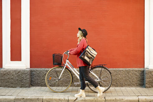 Young woman wearing hat looking away while walking with bicycle against red wall - JSMF01933