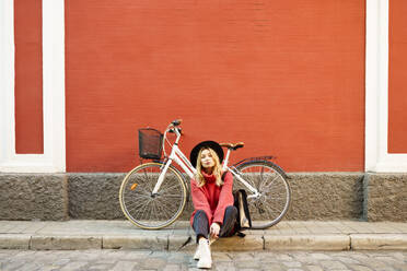 Blonde Frau mit Hut, die auf dem Gehweg an einem Fahrrad sitzt - JSMF01934