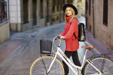 Woman wearing hat looking away while walking with bicycle on footpath in city - JSMF01945