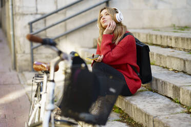 Thoughtful woman with headphones looking away while sitting on steps - JSMF01948