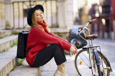 Young blond woman staring while sitting on steps by bicycle - JSMF01954