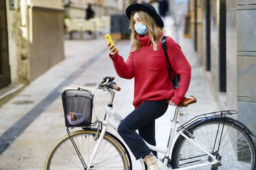 Junge Frau mit Gesichtsmaske, die ein Mobiltelefon benutzt, während sie mit dem Fahrrad auf dem Gehweg sitzt - JSMF01957