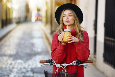 Blond woman wearing hat drinking juice while standing with bicycle on footpath - JSMF01961