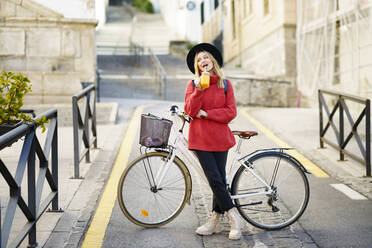 Young woman wearing hat drinking juice while standing with bicycle on road in city - JSMF01965