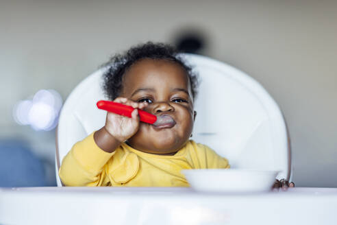 Baby girl eating with spoon while sitting on high chair at home - OCMF02016