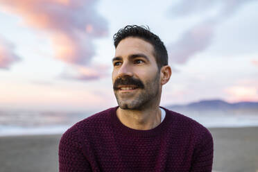 Hübscher Mann in kastanienbraunem Pullover träumt am Strand bei Sonnenaufgang - MIMFF00547