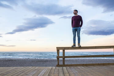 Man with hands in pockets standing on wooden railing at beach against sky - MIMFF00548