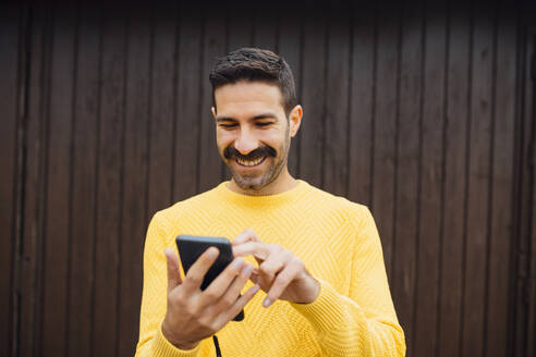 Smiling mid adult man using mobile phone against wooden wall - MIMFF00554