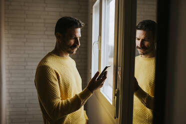 Seriöser Mann, der sein Smartphone benutzt, während er zu Hause am Fenster steht - MIMFF00555