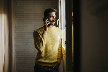 Mittlerer Erwachsener, der durch ein Fenster schaut, während er zu Hause mit seinem Handy telefoniert - MIMFF00556