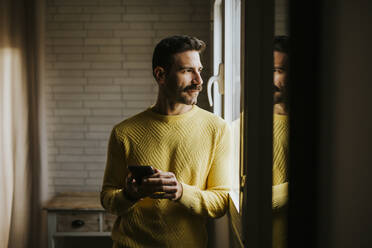 Nachdenklicher Mann, der durch ein Fenster schaut, während er zu Hause ein Mobiltelefon in der Hand hält - MIMFF00557