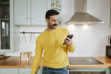 Gutaussehender Mann in gelbem Pullover, der sein Mobiltelefon an der Küchentheke benutzt - MIMFF00559