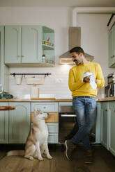 Mid adult man drying hands while looking at Siberian Husky dog in kitchen - MIMFF00561