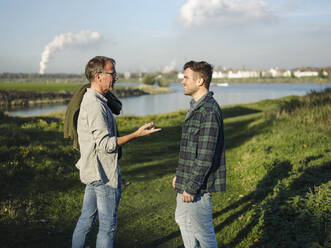 Father talking with son while standing on grass at riverbank - GUSF05169