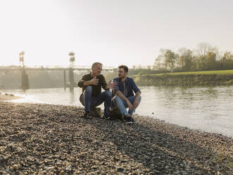 Father and son talking with each other while sitting on rock at riverbank during autumn - GUSF05173