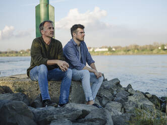 Thoughtful father and son looking away while sitting on rock against sky - GUSF05176