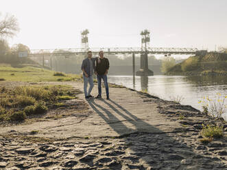 Son and father standing at riverbank during autumn - GUSF05180