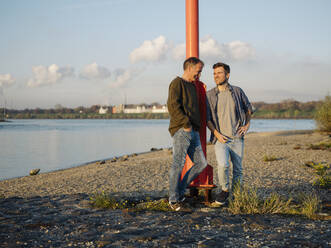 Smiling son and father leaning by pole while standing at riverbank - GUSF05182