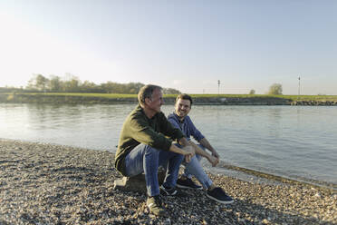 Father and son spending leisure time sitting at riverbank against clear sky - GUSF05184