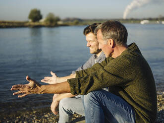 Vater gestikuliert im Gespräch mit seinem Sohn am See an einem sonnigen Tag - GUSF05186