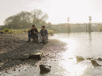 Father and son discussing at riverbank on sunny day - GUSF05189